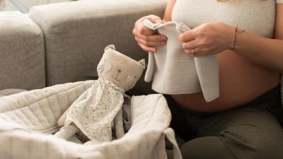 Těhotná žena drží oblečení pro miminko a vedle sebe má výbavičku.