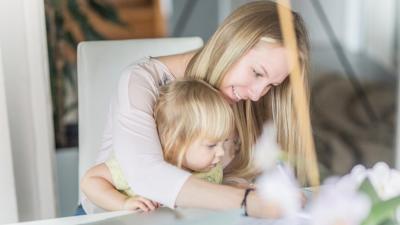 Maminka s dítětem v náruči píše na desce pracovního stolu