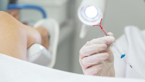 Doktor drží v ruce nitroděložní tělísko a chystá se ho zavést žene na gynekologickém křeslu.