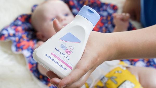 Maminka drží dětský olejíček sebamed Baby v ruce, v pozadí leží usměvavé batole