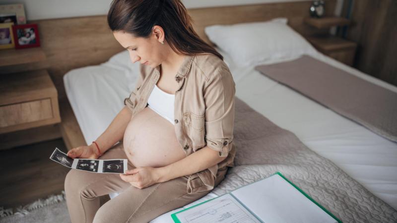 Těhotná žena sedí na posteli a prohlíží si fotky z ultrazvuku, screening ve druhém trimestru.