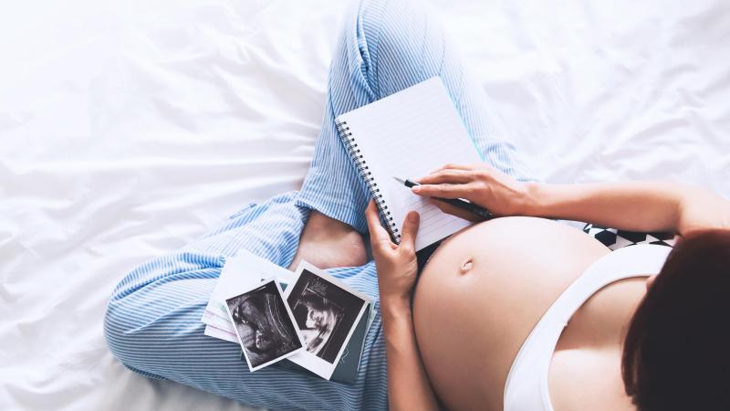 Těhotná žena sedí na posteli a prohlíží si fotky z ultrazvuku, screening ve druhém trimestru.
