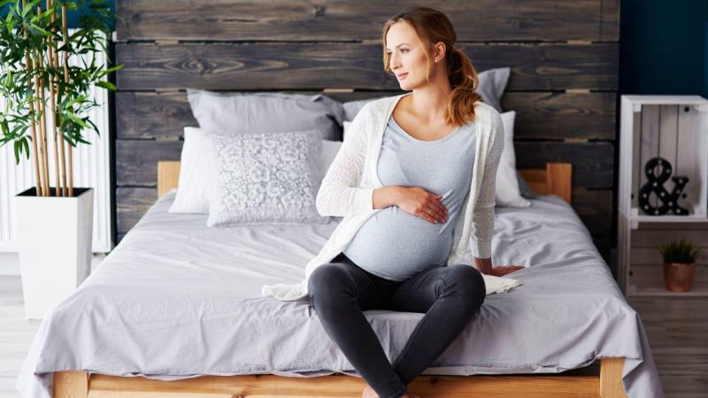 Těhotná žena sedí na posteli a drží se za břicho