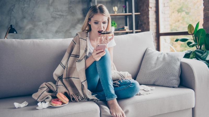 Žena na obrázku jí donuty a dívá se na mobilní telefon.