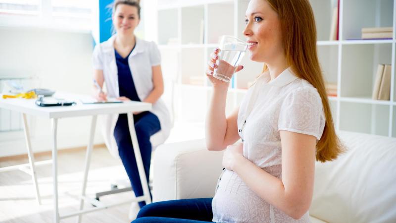 Těhotná žena u lékařky pije čistou vodu. Test na těhotenskou cukrovku.