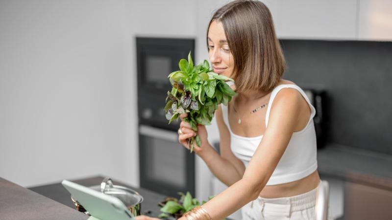 Žena drží v ruce bylinky a hledá recept na přípravu vaginální napářky v kuchyni.