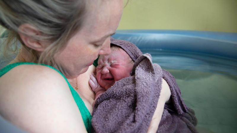 Maminka drží novorozence v bazénku po domácím porodu