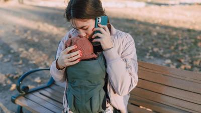 Žena líbá dítě v nosítku na temeno hlavy a zároveň volá z mobilního telefonu.
