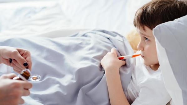 Nemocné dítě leží v posteli s teploměrem v puse, maminka mu podává léky. Čepy na mandlích.