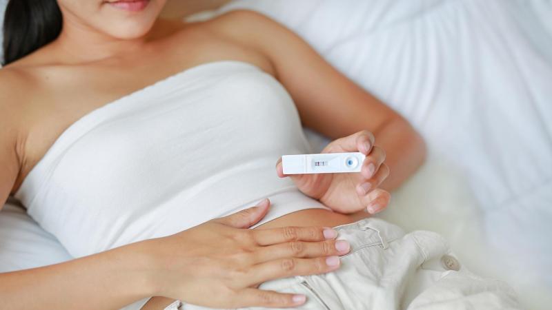 žena leží na posteli a drží v ruce pozitivní těhotenský test.